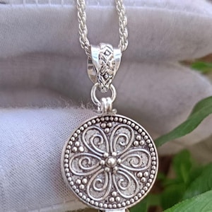 May include: A silver-colored pendant necklace featuring a round, detailed locket. The locket has a floral design with a beaded border and is suspended from a delicate chain. The pendant is approximately 1 inch in diameter.