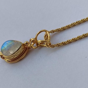 May include: A gold-colored necklace with a pendant featuring a pear-shaped, iridescent gemstone. The pendant is set in a decorative gold bezel and hangs from a delicate chain. The necklace is displayed on a white surface.