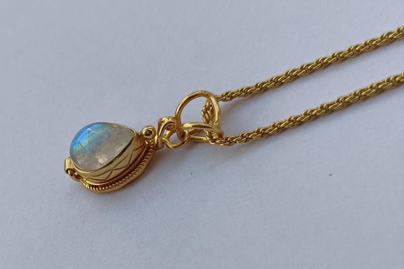 May include: A gold-colored necklace with a pendant featuring a pear-shaped, iridescent gemstone. The pendant is set in a decorative gold bezel and hangs from a delicate chain. The necklace is displayed on a white surface.