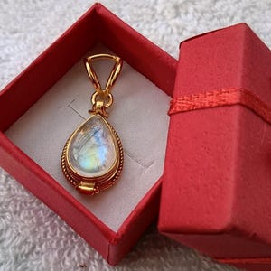 May include: A gold-colored pendant with a pear-shaped, iridescent gemstone is displayed inside a red gift box. The pendant has a detailed, rope-like border and a simple bail. The box is open, revealing the pendant against a white interior.