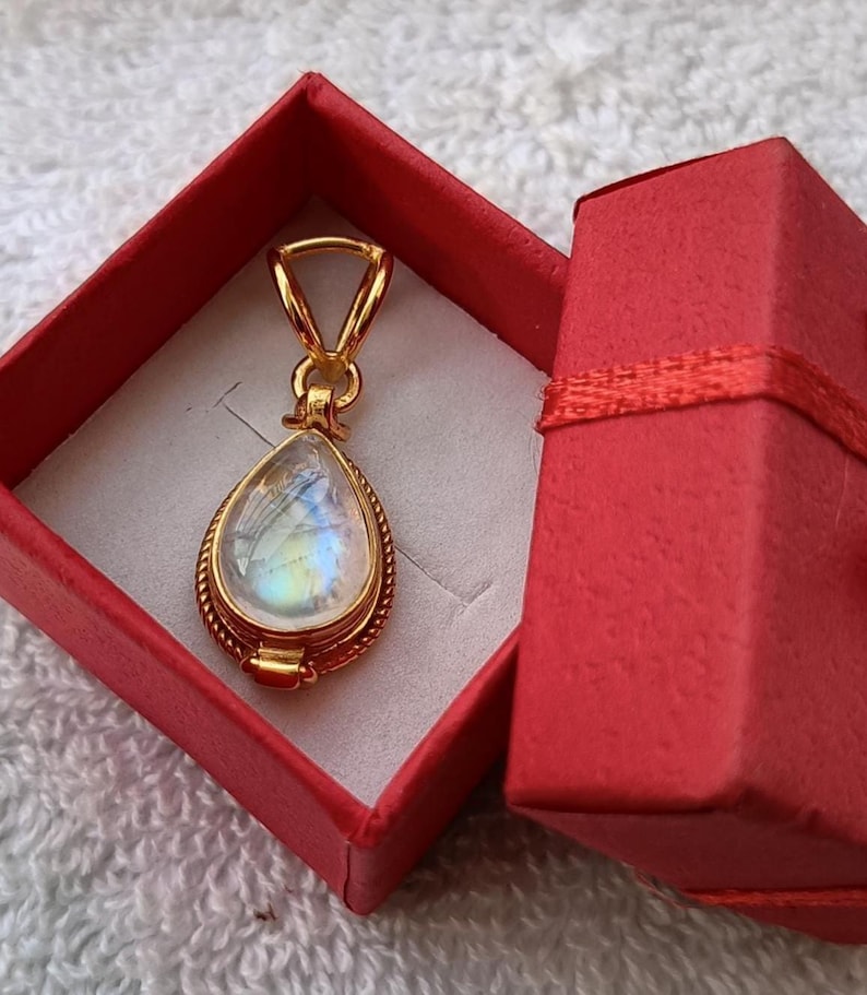 May include: A gold-colored pendant with a pear-shaped, iridescent gemstone is displayed inside a red gift box. The pendant has a detailed, rope-like border and a simple bail. The box is open, revealing the pendant against a white interior.