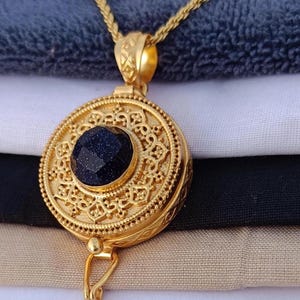 May include: A gold-colored pendant necklace with a round, ornate locket design. The locket features a dark blue, faceted stone in the center and intricate detailing around the edges. The necklace is displayed against a backdrop of folded fabric.