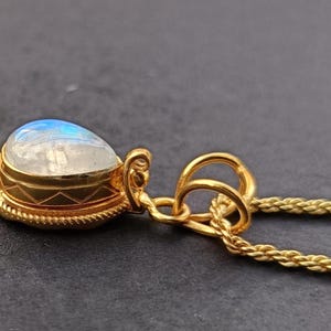 May include: A gold-colored pendant necklace featuring a teardrop-shaped moonstone. The moonstone has a blue and white iridescent sheen. The pendant is encased in a gold setting with decorative detailing and hangs from a delicate gold chain.