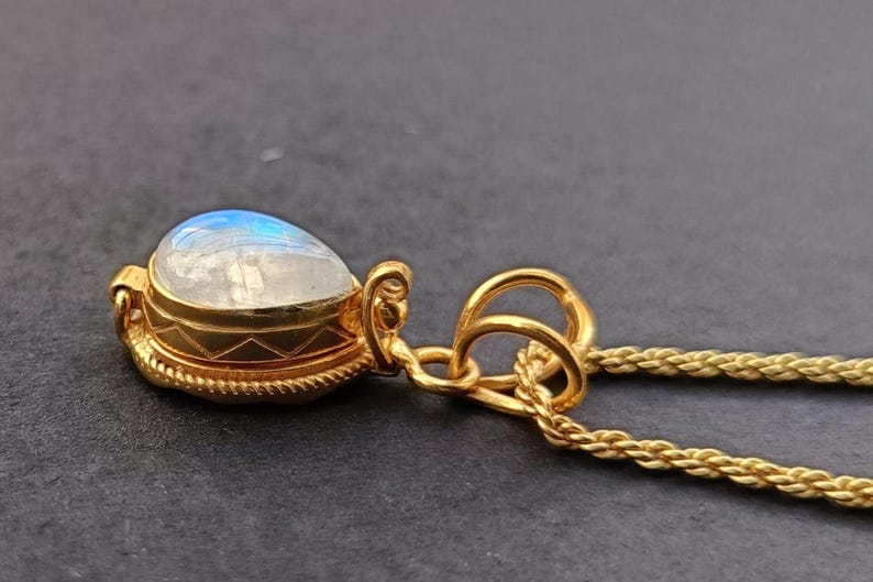 May include: A gold-colored pendant necklace featuring a teardrop-shaped moonstone. The moonstone has a blue and white iridescent sheen. The pendant is encased in a gold setting with decorative detailing and hangs from a delicate gold chain.
