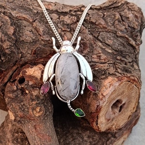 May include: Silver necklace with a beetle pendant featuring a clear quartz stone with black inclusions, accented with red and green gemstones.