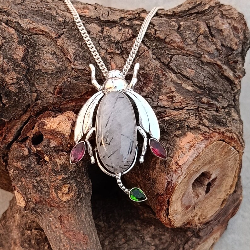 May include: Silver necklace with a beetle pendant featuring a clear quartz stone with black inclusions, accented with red and green gemstones.