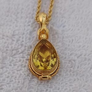 May include: A gold-colored pendant necklace with a pear-shaped, faceted yellow gemstone. The pendant is suspended from a delicate chain. The gemstone is set in a gold bezel with a rope-like border.