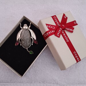 May include: A silver beetle brooch with a clear quartz stone and red and green gemstones. The brooch is in a white box with a red ribbon and the text "Crystal Jewelry" printed on it.