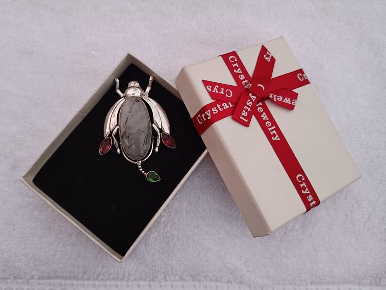 May include: A silver beetle brooch with a clear quartz stone and red and green gemstones. The brooch is in a white box with a red ribbon and the text "Crystal Jewelry" printed on it.