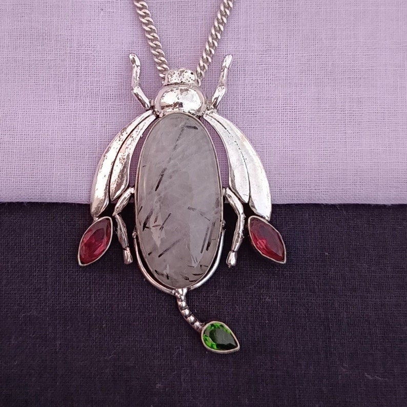May include: Silver pendant necklace featuring a clear quartz stone with black inclusions, set in a silver bug design with red and green gemstone accents.
