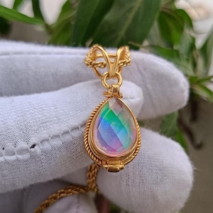 May include: A gold-colored pendant necklace featuring a teardrop-shaped iridescent gemstone. The gemstone displays a spectrum of colors, including pink, green, and blue. The pendant is encased in a gold-toned setting with a rope-like border and hangs from a delicate gold chain.