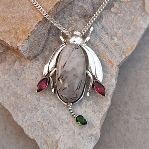 May include: Silver pendant necklace featuring a clear quartz stone with black inclusions, set in a beetle design with red and green accents.