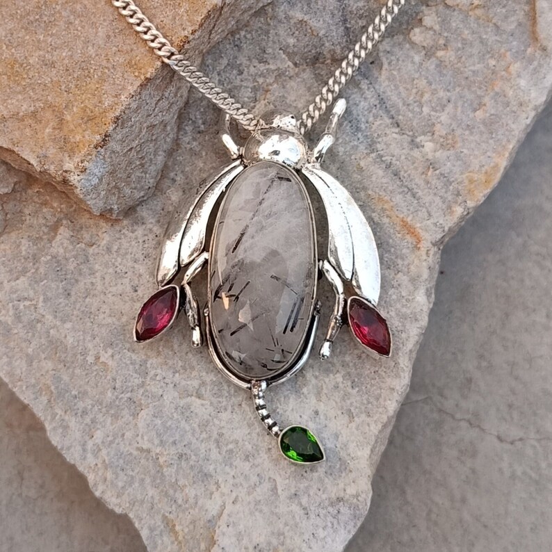 May include: Silver pendant necklace featuring a clear quartz stone with black inclusions, set in a beetle design with red and green accents.