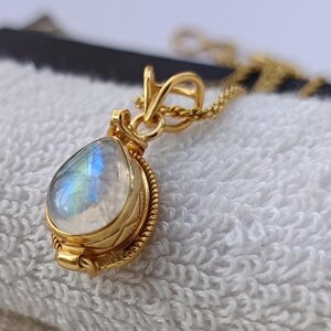 May include: A gold pendant necklace featuring a teardrop-shaped moonstone. The moonstone displays iridescent blue and white hues. The pendant is set in a gold bezel with a decorative rope-like border, suspended from a delicate gold chain.