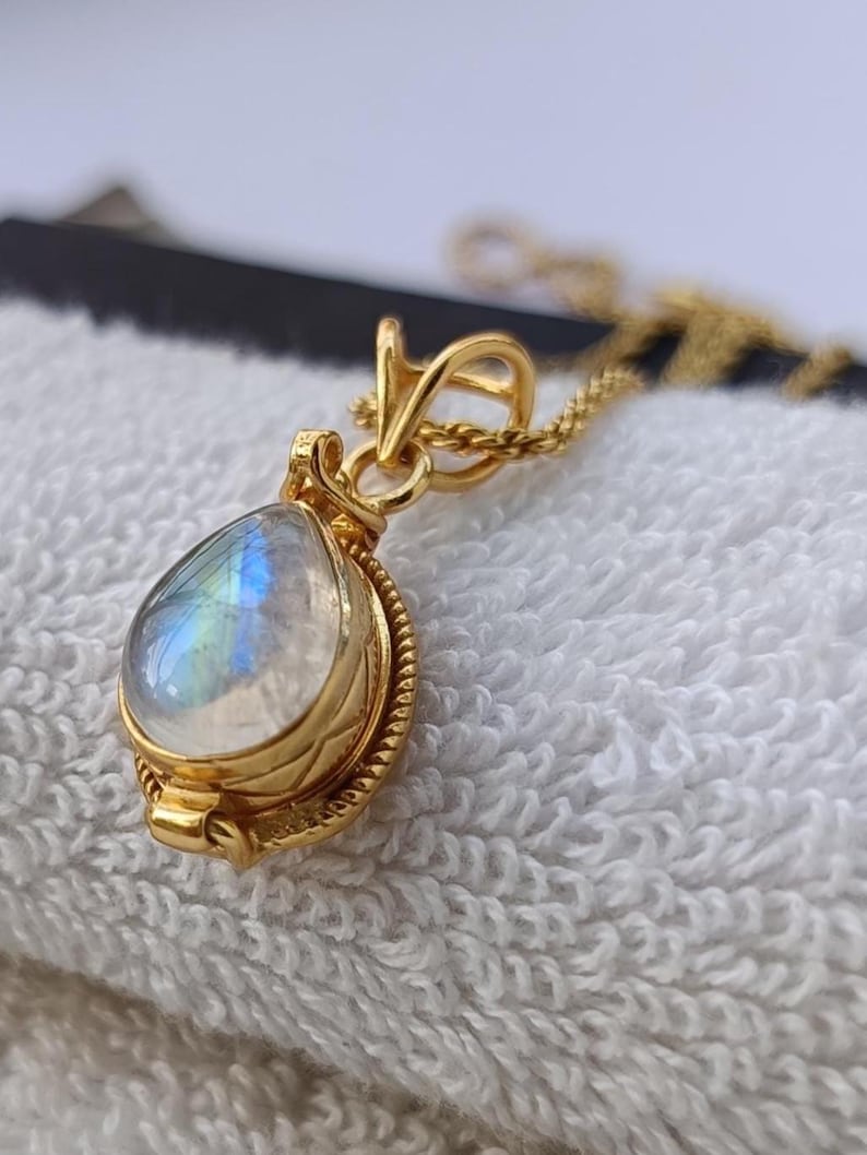 May include: A gold pendant necklace featuring a teardrop-shaped moonstone. The moonstone displays iridescent blue and white hues. The pendant is set in a gold bezel with a decorative rope-like border, suspended from a delicate gold chain.