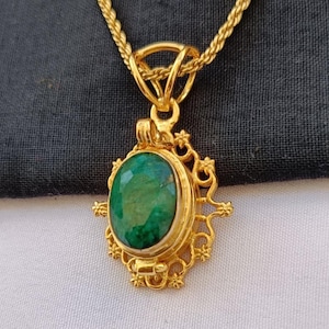 May include: A gold-colored pendant necklace featuring an oval, faceted emerald gemstone. The emerald is set in a decorative, filigree-style frame. The necklace chain is a twisted rope design.