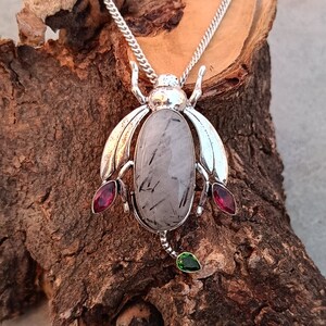 May include: A silver pendant necklace featuring a beetle design with a clear quartz stone and red and green accents.