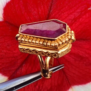 May include: A gold ring with a large, purple gemstone set in a coffin-shaped setting. The ring is detailed with a beaded border and intricate designs.