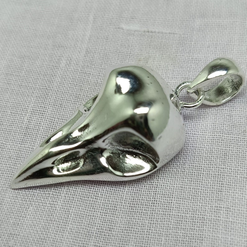 Puede incluir: Un colgante de plata con forma de cr&aacute;neo de p&aacute;jaro con un bucle para sujetar una cadena.