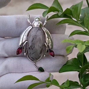 May include: Silver pendant featuring a grey and black gemstone with a silver insect design. The insect has red and green accents.