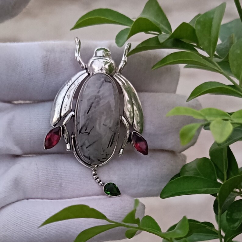 May include: Silver pendant featuring a grey and black gemstone with a silver insect design. The insect has red and green accents.