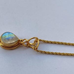 May include: A gold-colored necklace with a teardrop-shaped pendant. The pendant features a pearlescent, iridescent stone set in a gold bezel. The chain is a delicate, woven design. The necklace is displayed on a white surface.