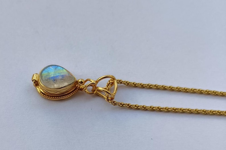 May include: A gold-colored necklace with a teardrop-shaped pendant. The pendant features a pearlescent, iridescent stone set in a gold bezel. The chain is a delicate, woven design. The necklace is displayed on a white surface.