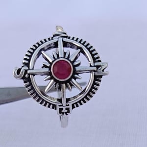May include: A silver-colored ring featuring a compass design. The ring has a central red gemstone and a detailed, textured compass rose. The ring's design includes directional markers and a circular frame.