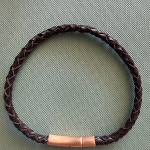 May include: A dark brown, braided leather bracelet with a copper-coloured clasp. The bracelet is circular and made of interwoven leather strips. The clasp is rectangular and connects the two ends of the bracelet.
