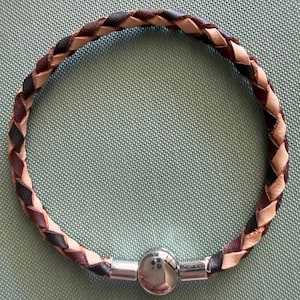 May include: A braided leather bracelet in shades of brown, black, and tan. The bracelet has a silver-coloured, spherical clasp. The bracelet is circular and is shown on a light green surface. The bracelet's diameter is approximately 6 cm.