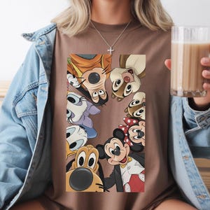 May include: A brown t-shirt with a graphic of Disney characters such as Mickey Mouse, Minnie Mouse, Goofy, and Chip and Dale. The characters are arranged in a circle. The person is wearing a denim jacket.