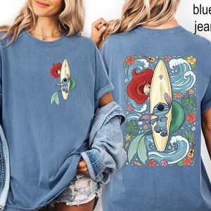 May include: Blue jean-colored t-shirt featuring a cartoon design of Ariel and Stitch surfing on a yellow surfboard. The front of the shirt shows a smaller version of the design. The back of the shirt has a larger design with waves, flowers, and tropical elements.
