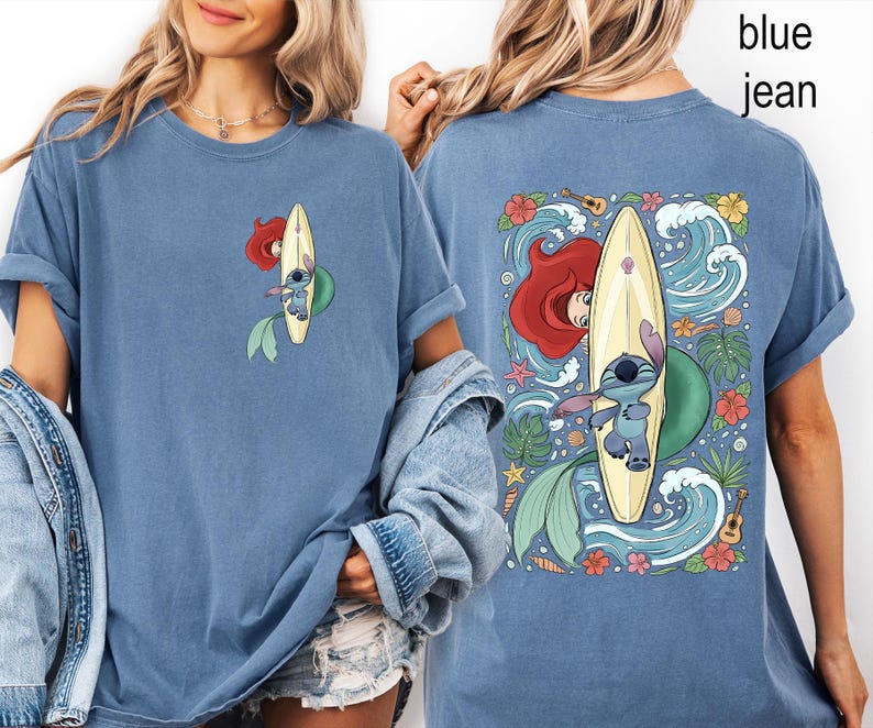 May include: Blue jean-colored t-shirt featuring a cartoon design of Ariel and Stitch surfing on a yellow surfboard. The front of the shirt shows a smaller version of the design. The back of the shirt has a larger design with waves, flowers, and tropical elements.
