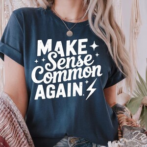 May include: Navy blue t-shirt with the white text "MAKE Sense Common AGAIN" and a lightning bolt graphic. The shirt has rolled-up sleeves and a relaxed fit. The design includes star accents.