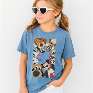 May include: A pale blue t-shirt with a vibrant graphic of cartoon characters such as Mickey Mouse, Goofy, and Chip and Dale. The shirt is made of a soft material, suitable for casual wear.