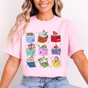 May include: Pink t-shirt featuring a collection of illustrated books with various cartoon characters. Each book has a different color and design, with characters like a monkey, a frog, and a rooster.