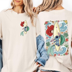 May include: Cream-colored t-shirt featuring a cartoon design of Ariel and Stitch surfing on a yellow surfboard. The front has a small image, while the back showcases a larger, detailed illustration with waves, flowers, and a ukulele.
