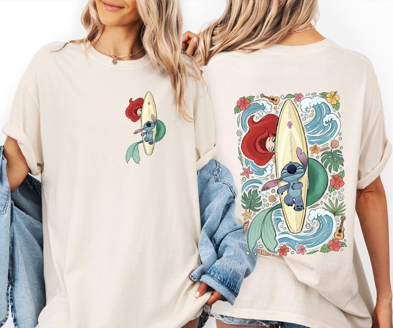 May include: Cream-colored t-shirt featuring a cartoon design of Ariel and Stitch surfing on a yellow surfboard. The front has a small image, while the back showcases a larger, detailed illustration with waves, flowers, and a ukulele.