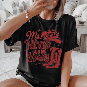 May include: Black t-shirt with a red graphic featuring a cowboy hat and boots, along with the text "Ms. Never Do No Wrong." The design includes stars and decorative elements, creating a Western-themed aesthetic.