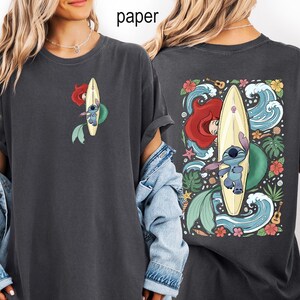 May include: Dark gray t-shirt featuring a cartoon design of Ariel and Stitch surfing. The front has a small image of Ariel and Stitch on a surfboard. The back has a larger image with waves, flowers, and tropical leaves.