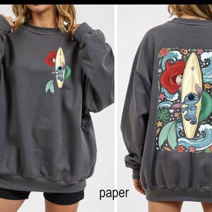 May include: Dark gray sweatshirt featuring a front and back graphic. The front graphic shows Ariel and Stitch on a surfboard. The back graphic shows Ariel, Stitch, waves, flowers, and a ukulele. The word "paper" is printed at the bottom.