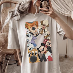 May include: A pale grey t-shirt with a vibrant graphic of Disney cartoon characters, such as Mickey Mouse, Goofy, and Donald Duck. The characters are arranged in a circular design. The shirt has short sleeves and a loose fit.