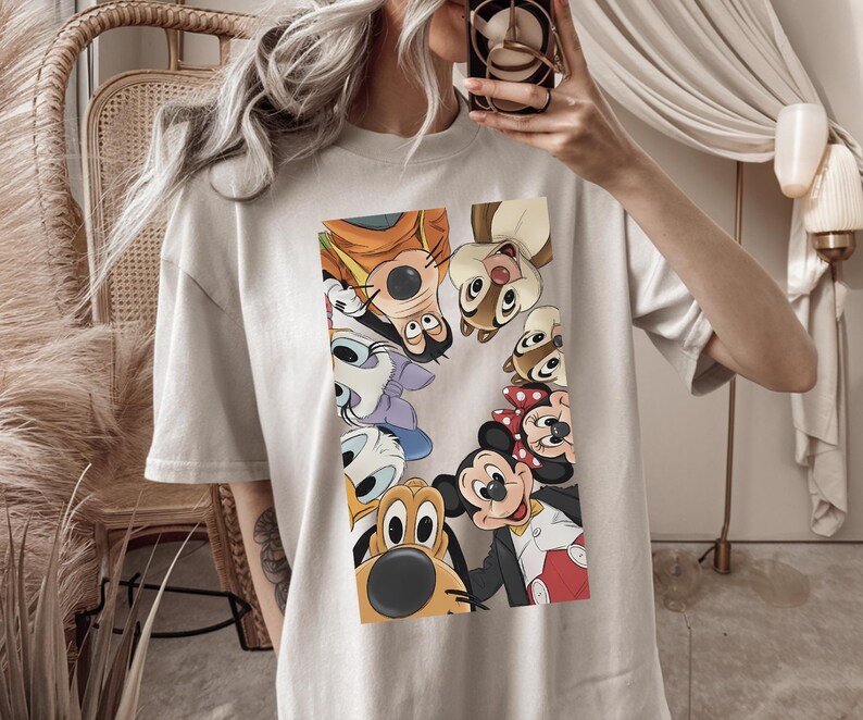 May include: A pale grey t-shirt with a vibrant graphic of Disney cartoon characters, such as Mickey Mouse, Goofy, and Donald Duck. The characters are arranged in a circular design. The shirt has short sleeves and a loose fit.