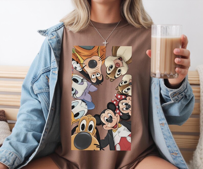 May include: A brown t-shirt with a graphic of Disney characters such as Mickey Mouse, Minnie Mouse, Goofy, and Chip and Dale. The characters are arranged in a circle. The person is wearing a denim jacket.