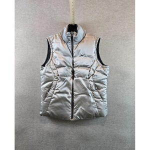 May include: Silver puffer vest with a black zipper and collar. Features a small Nike logo on the chest. The vest has a quilted design and two side pockets. A stylish and warm outerwear option.