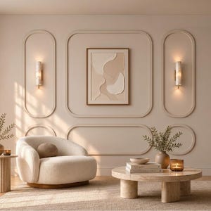 May include: A cream-colored armchair with a rounded back sits beside a fluted side table holding a vase with greenery. A low, cylindrical stone coffee table features books, a bowl, and a candle. The wall has beige molding and two sconces.