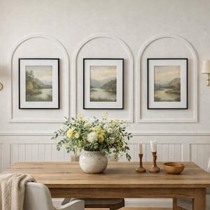 May include: Three framed landscape prints featuring muted green and brown scenery, displayed within white arched wall moldings. A wooden dining table sits below, holding a ceramic vase filled with yellow and white flowers and two candles.