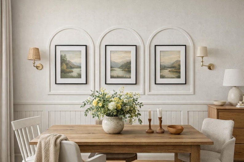 May include: Three framed landscape prints featuring muted green and brown scenery, displayed within white arched wall moldings. A wooden dining table sits below, holding a ceramic vase filled with yellow and white flowers and two candles.
