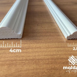May include: Two white decorative molding pieces. One is 4 cm wide, the other is 2.5 cm wide. The molding has a curved, decorative edge. The background is a wooden surface. The logo "moldart wall molding" is in the bottom right corner.