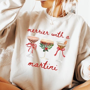 May include: A sand-colored sweatshirt with the text "merrier with a martini" in red script. The graphic features three martini glasses with festive decorations. The person in the image is holding a black mug.